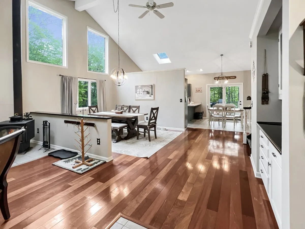 A spacious, sun-drenched dining and living area with large windows revealing scenic views. The rustic dining table is set for a gathering, and the open layout connects seamlessly to the living space, which features cozy seating and a stove, perfect for family meals or celebrations.
