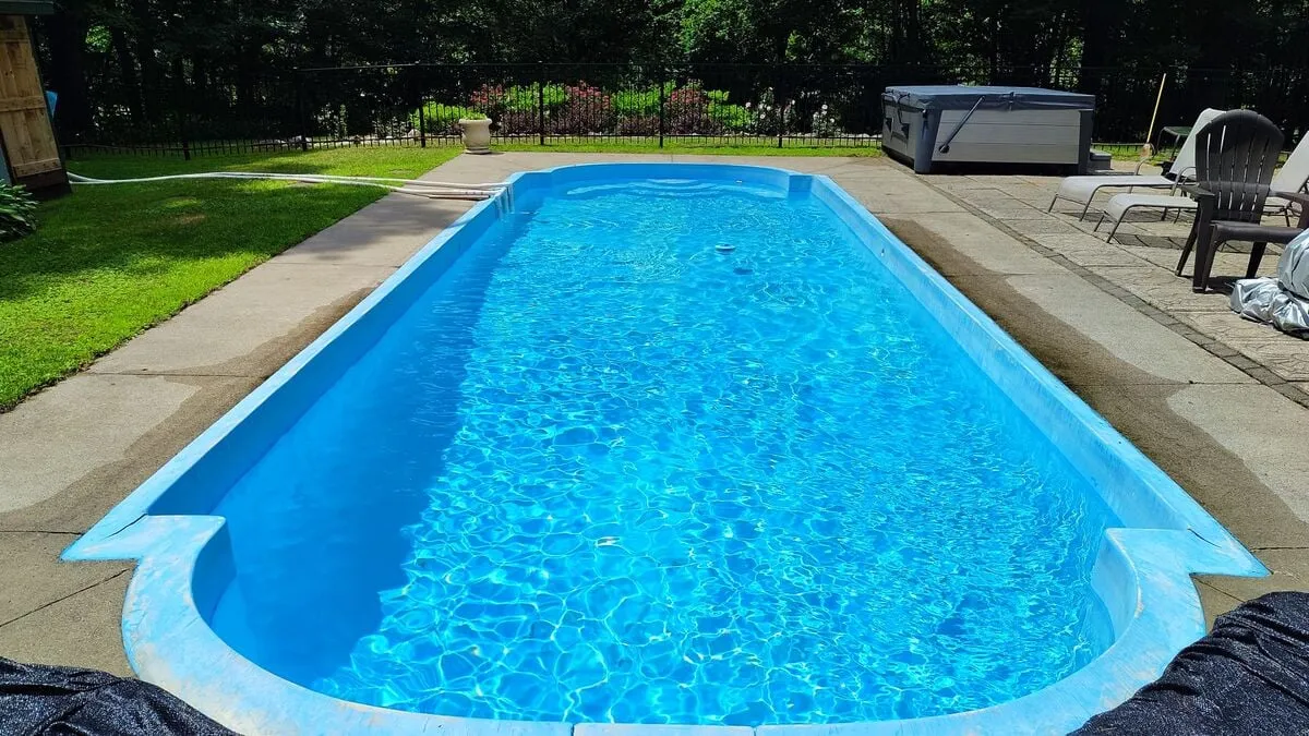 This inviting exterior image showcases a sparkling swimming pool surrounded by lush green grass. Lounge chairs line the poolside, and a bubbling hot tub sits adjacent to the pool, indicating spaces for sunbathing, swimming, and soaking. The scene is alive with the warm glow of sunset, suggesting festive gatherings and relaxation.