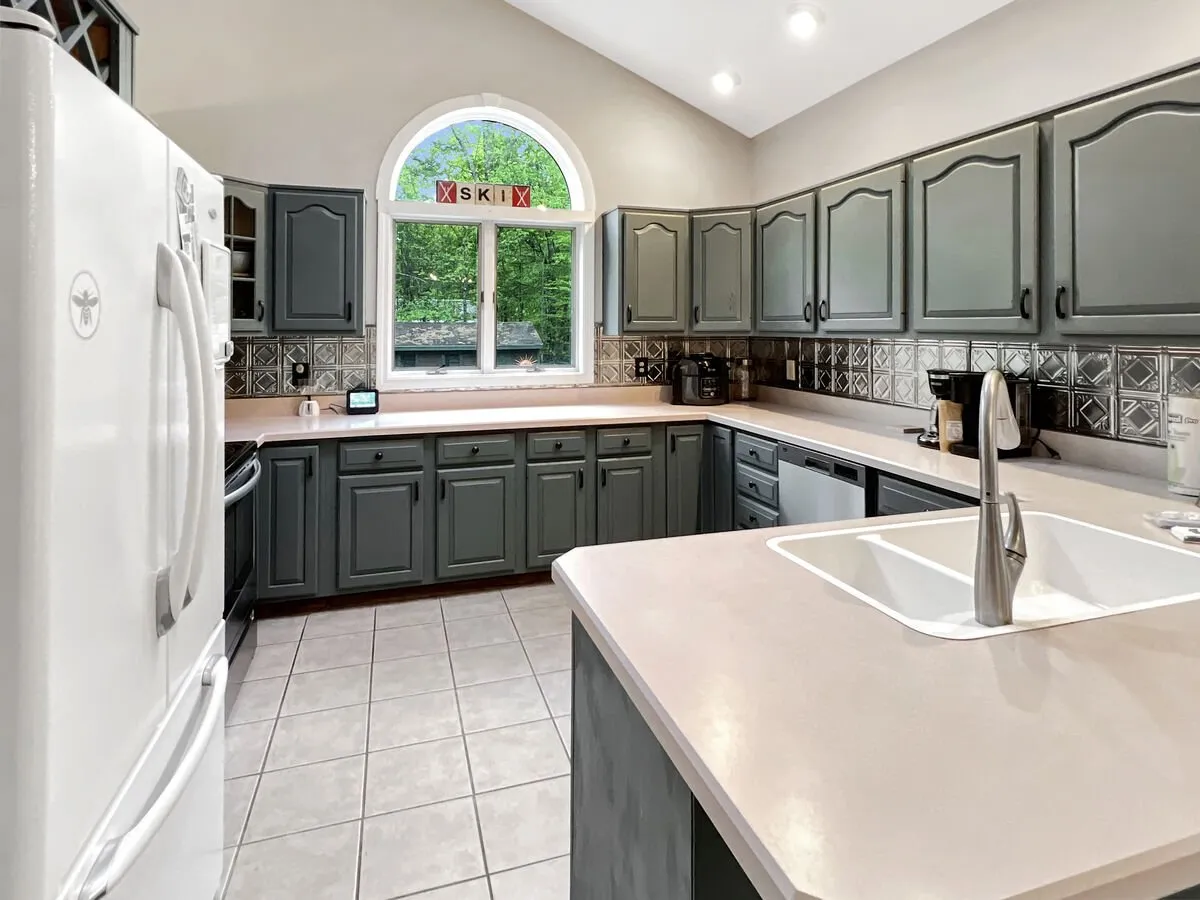This image centers on a bright, inviting kitchen with a spacious island, large window, and ample cabinetry. The setup is clearly designed for both everyday cooking and gathering, with the island providing a focal point for meal prep, socializing, or casual dining. Natural light floods the space, highlighting its modern yet cozy atmosphere.