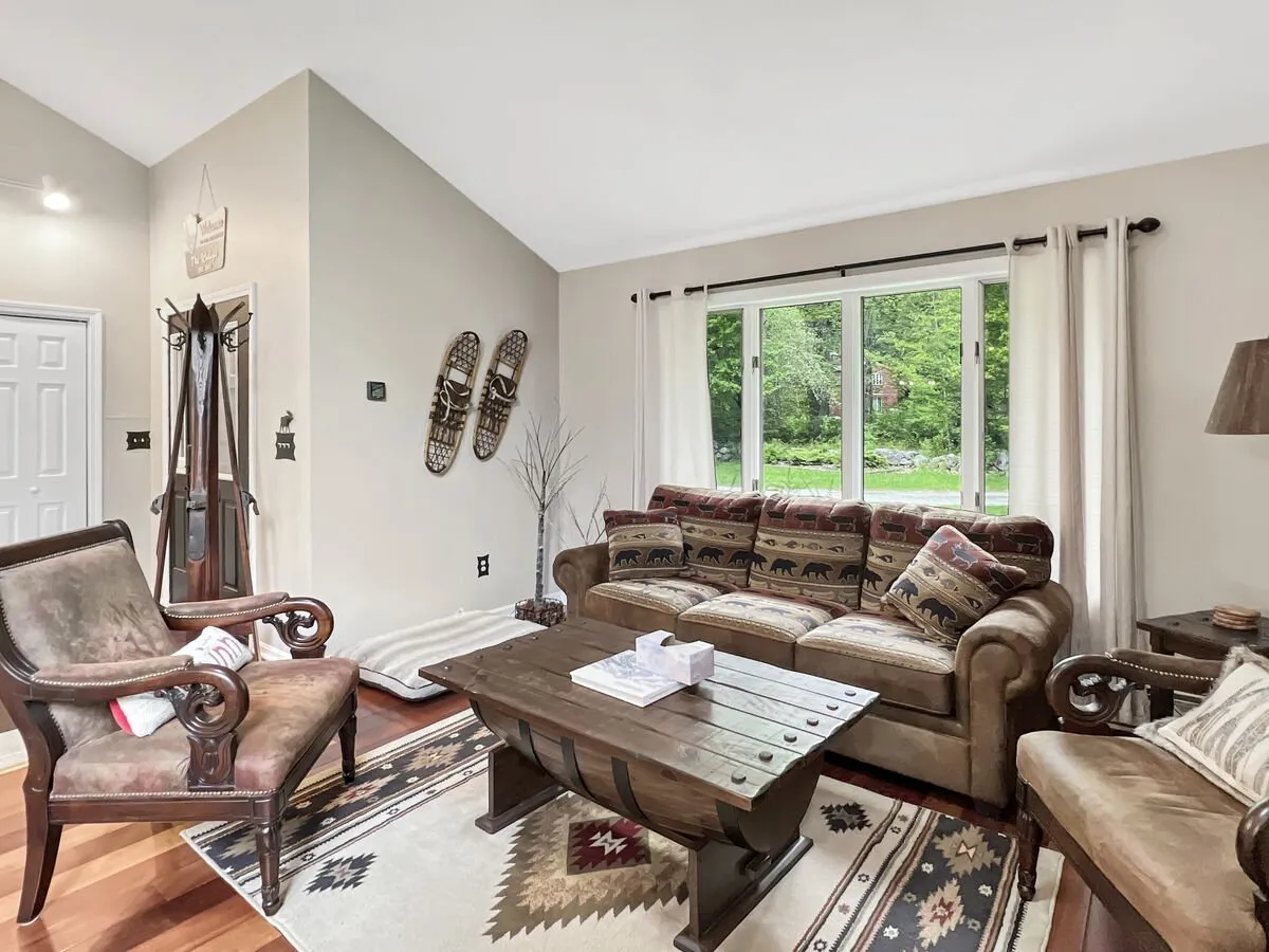 A cozy, sunlit living room with a comfortable sofa adorned with patterned cushions sits beside a large window that frames vibrant green forest views. A rustic wooden coffee table anchors the space, evoking a connection to the outdoors while providing an inviting spot to relax after a day of adventure.