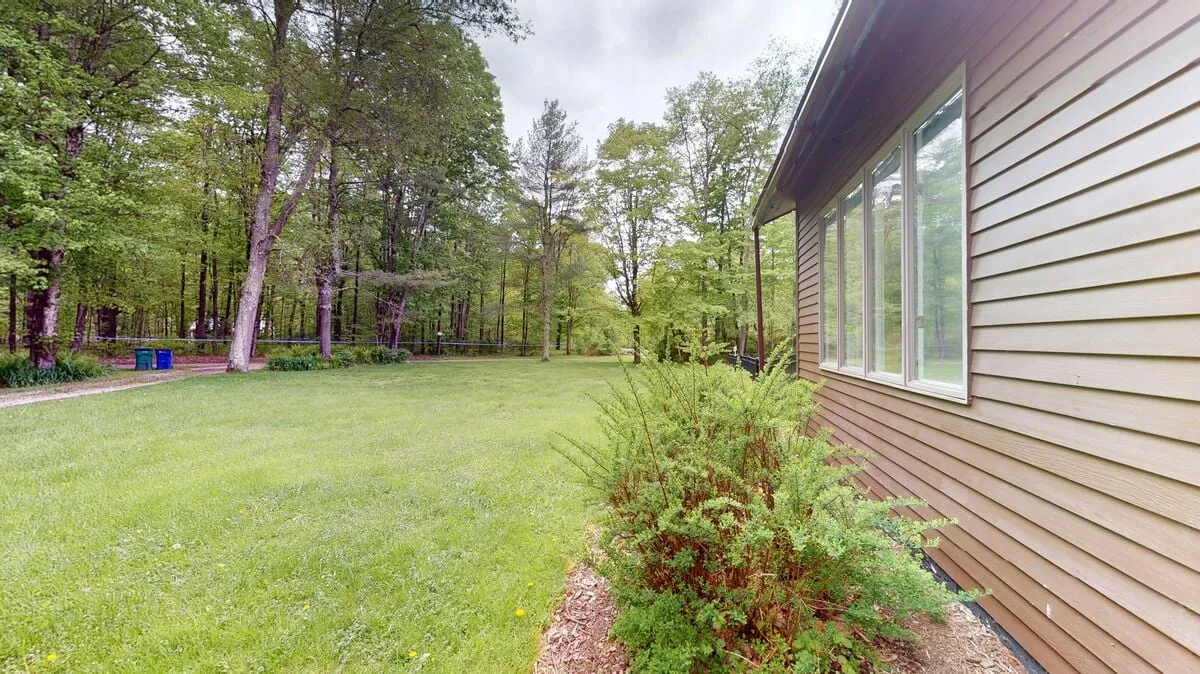 The scene shows a lush, green lawn bordered by a tree line and the side of the house, with bushes and open space inviting outdoor enjoyment. The tranquil mood suggests quiet mornings or evening gatherings after a day of exploration.