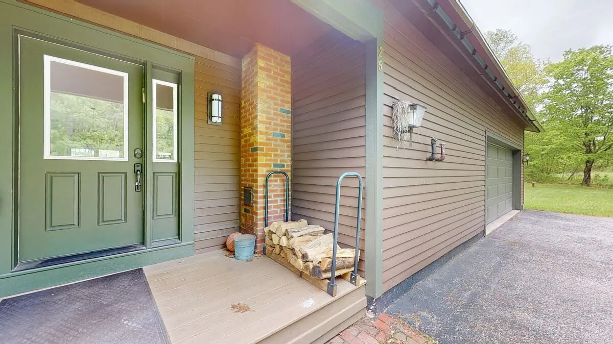 The front steps and porch feature a brick accent wall, wooden steps, and a stack of firewood, all framed by greenery. This cozy porch scene hints at gatherings, welcoming pets, and enjoying the outdoors in comfort.