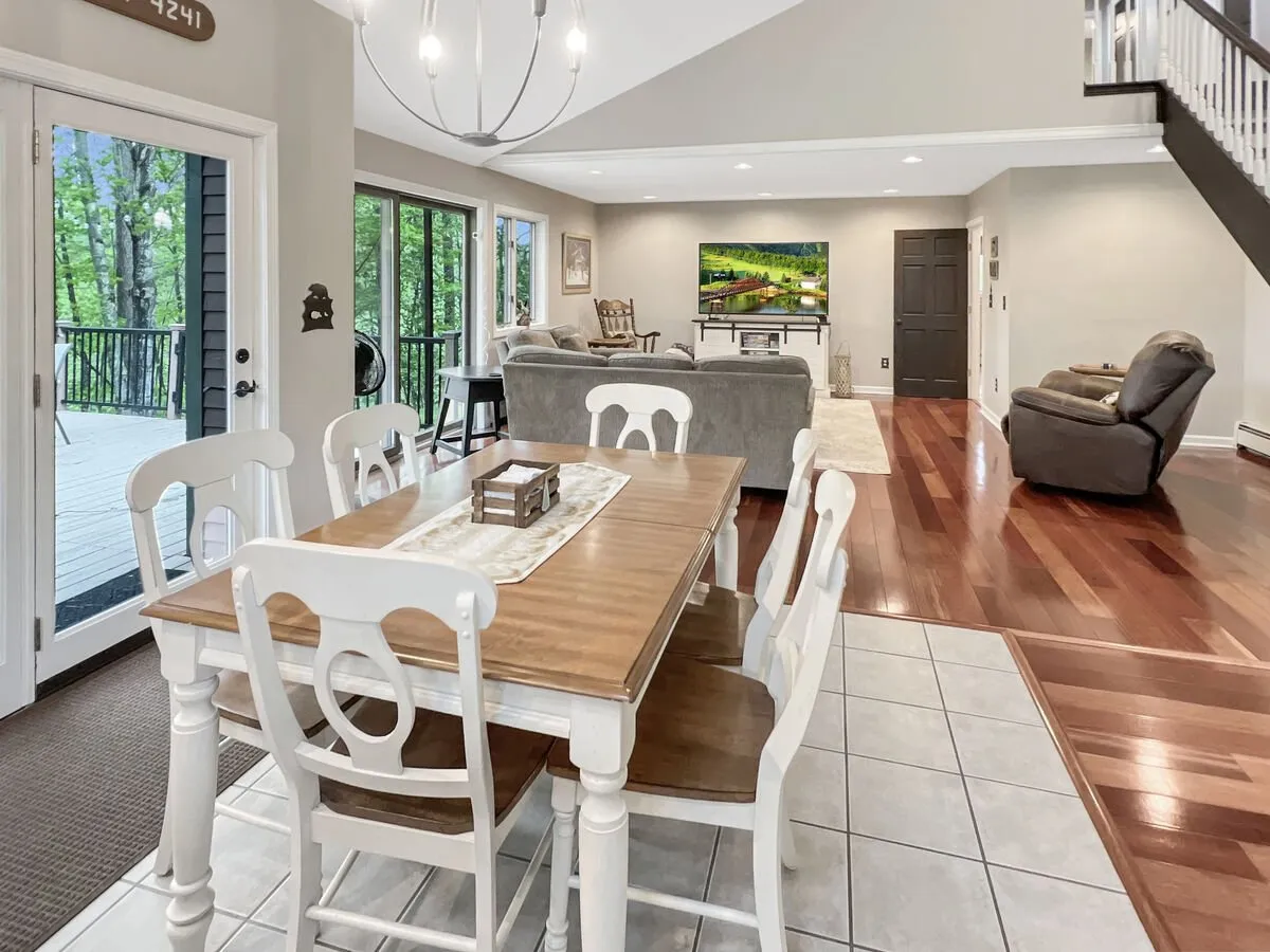 A warm, inviting area where the dining table and living room are integrated. Large windows let in abundant light, while the sofa and TV suggest spaces for both socializing and winding down. The wood floors gleam under natural light, enhancing the room's charm.