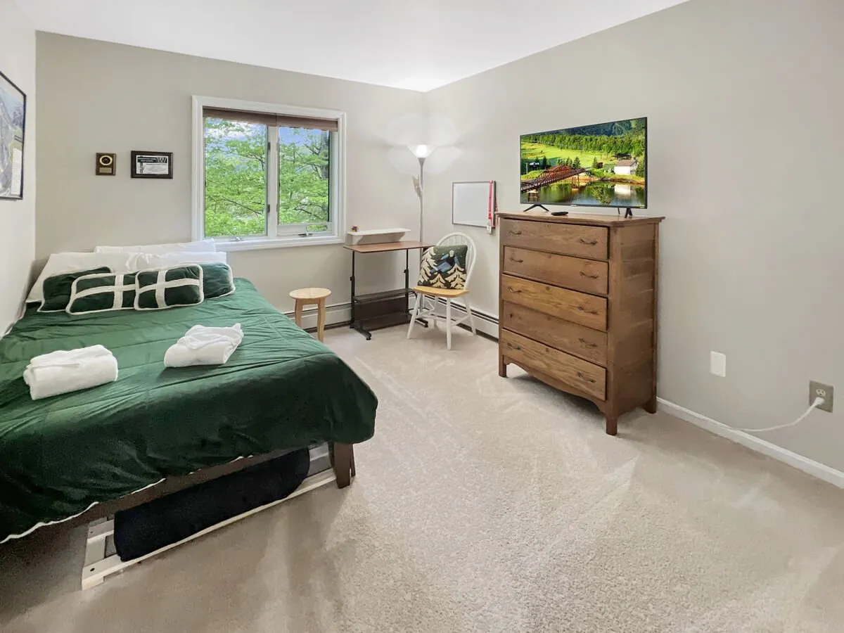 A bedroom featuring green bedding on the bed, a wooden dresser, a window with a view of green foliage, and a television showing a nature scene. The room feels relaxing and inviting.