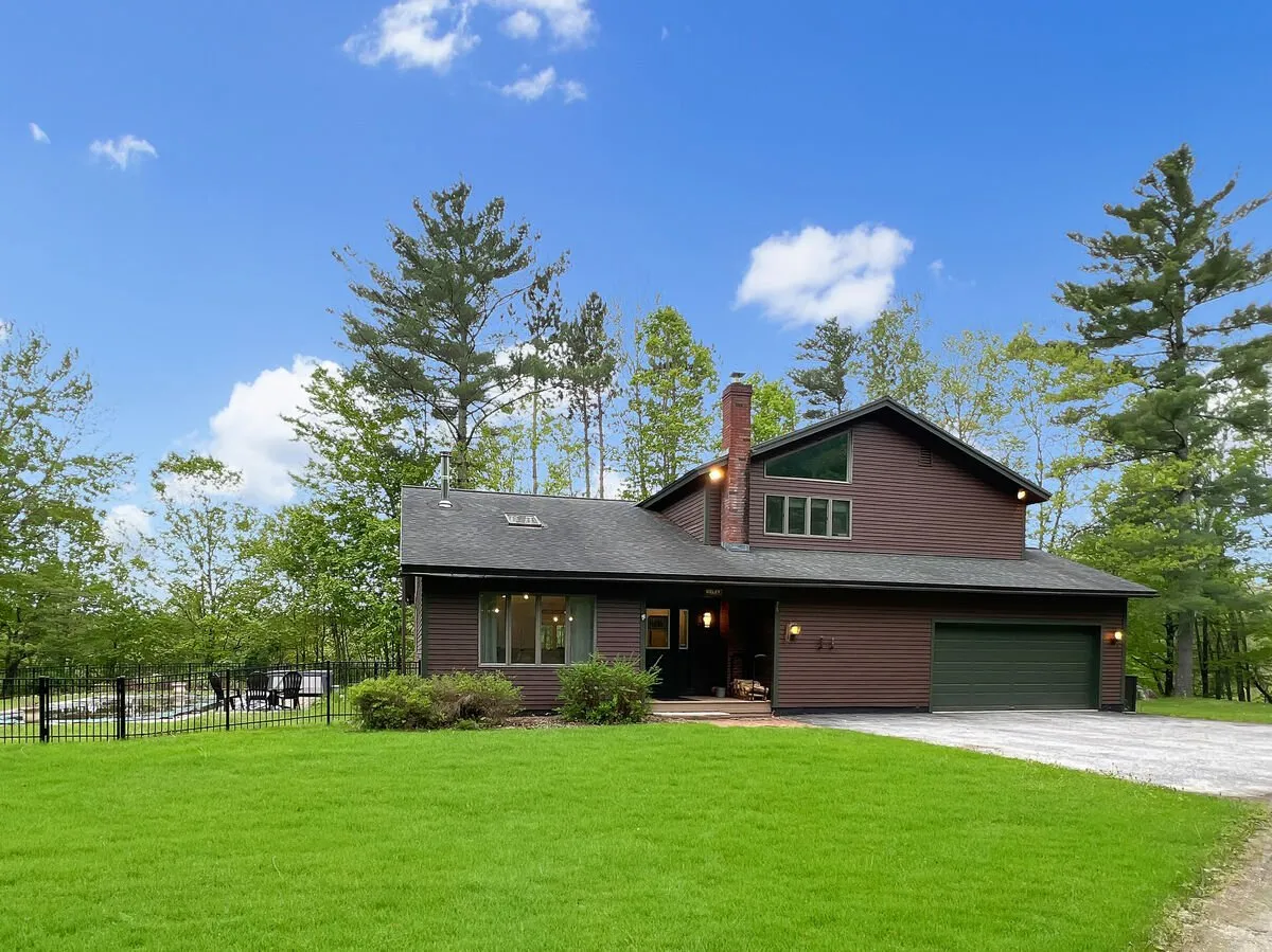 The image showcases the modern architecture of a spacious single-family home set against a backdrop of lush Vermont woods. The large front lawn is perfect for gatherings, and the inviting porch hints at relaxing evenings watching sunsets. Surrounding trees provide privacy and a sense of serene seclusion, making the scene feel peaceful and welcoming.