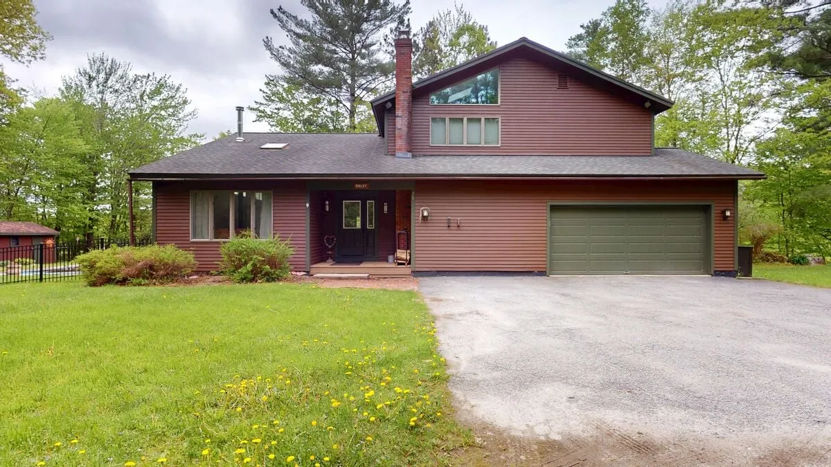 This exterior shot captures the main entrance of Sugar Hollow, flanked by large windows and a green garage door. The spacious yard and tranquil natural surroundings are visible, hinting at ample room for family gatherings and outdoor activities.