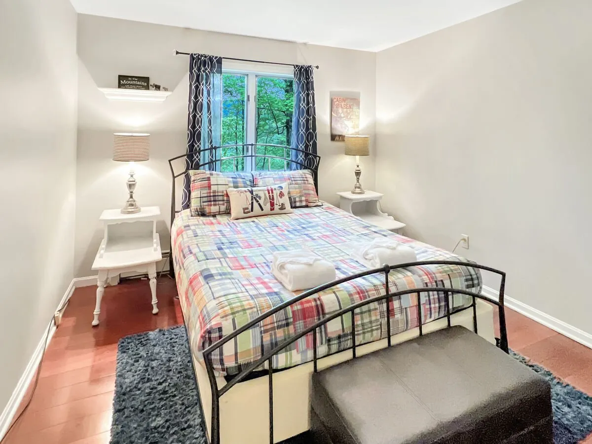 A cozy bedroom with plaid bedding and a window that frames lush greenery outside. A pair of nightstand lamps creates a soft, welcoming atmosphere. The nature views invite thoughts of waking up with your pet and heading straight outdoors for a morning walk.
