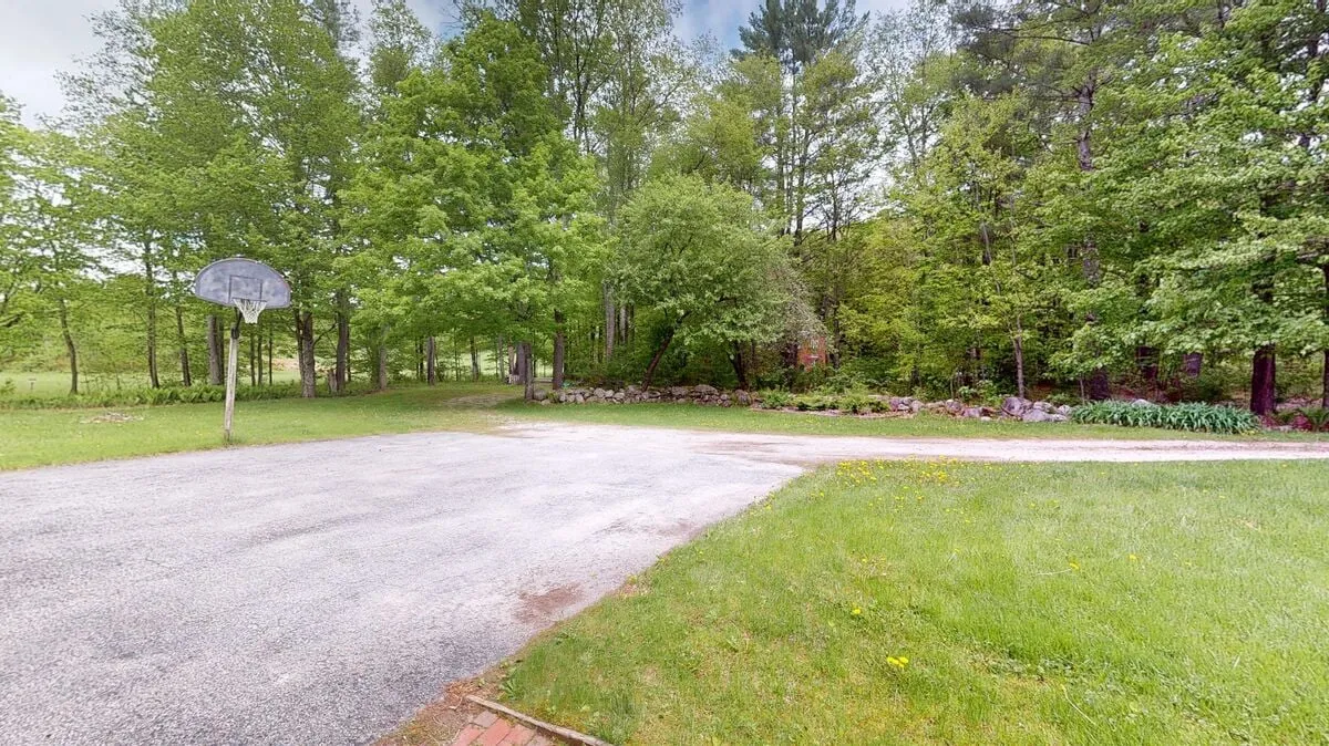 This image features a playful exterior scene with a basketball hoop, lush green lawn, and trees providing dappled shade. The expansive grassy area suggests room for picnics and outdoor games, while the surrounding woods evoke the proximity to hiking and biking trails.