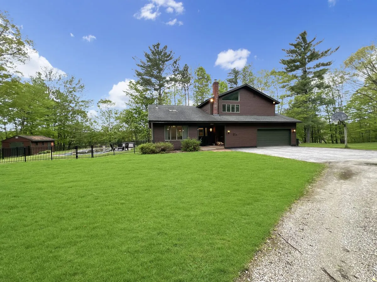 The image shows the house's facade surrounded by a well-manicured lawn, a basketball hoop for family fun, and mature trees that suggest a tranquil, natural setting. The porch offers a welcoming space, while the greenery alludes to the outdoor adventures in the Vermont countryside.